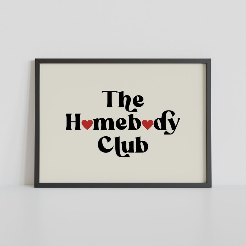 May include: Black and white print with the text "The Homebody Club" in a retro font. Two red hearts are placed between the words "Home" and "body".