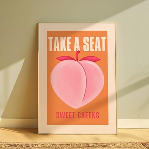 May include: A peach-colored poster with the text "Take a Seat" in white letters and "Sweet Cheeks" in pink letters. The poster features a stylized peach with a pink and red color scheme.
