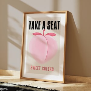 May include: A framed print with the text "Take a Seat Sweet Cheeks" in black and pink lettering. A pink peach illustration is in the center of the print.