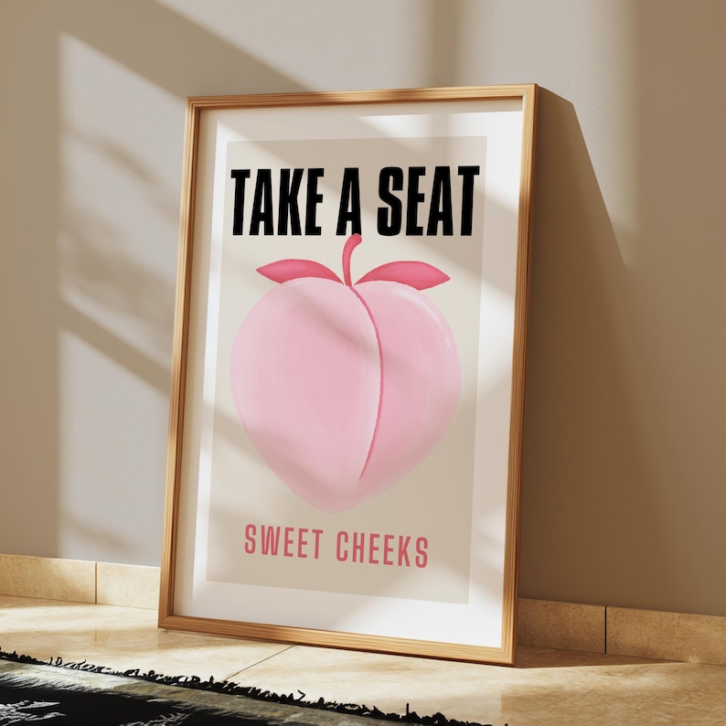 May include: A framed print with the text "Take a Seat Sweet Cheeks" in black and pink lettering. A pink peach illustration is in the center of the print.