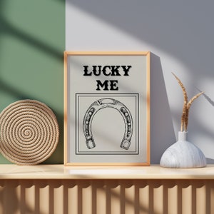 May include: A framed print with the text "LUCKY ME" and a black and white illustration of a horseshoe. The print is on a wooden shelf with a woven rope coaster and a white vase with dried grass.