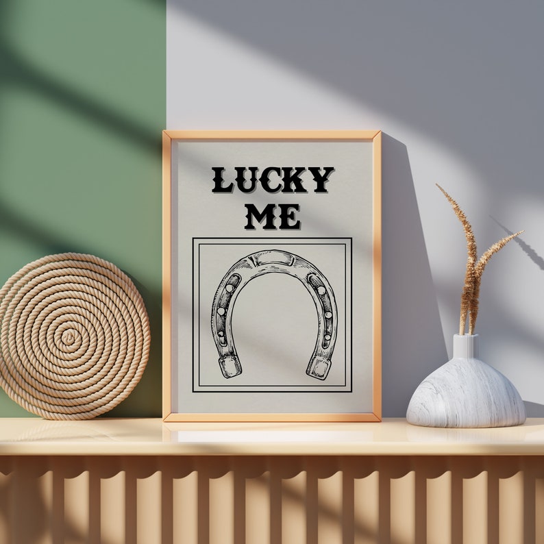 May include: A framed print with the text "LUCKY ME" and a black and white illustration of a horseshoe. The print is on a wooden shelf with a woven rope coaster and a white vase with dried grass.