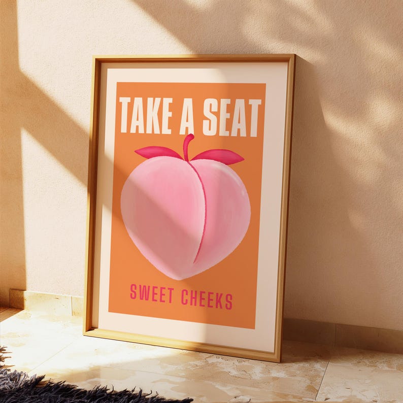 May include: A peach-colored poster with the text "Take a Seat Sweet Cheeks" in white and pink lettering. A peach with a pink blush is depicted in the center of the poster.