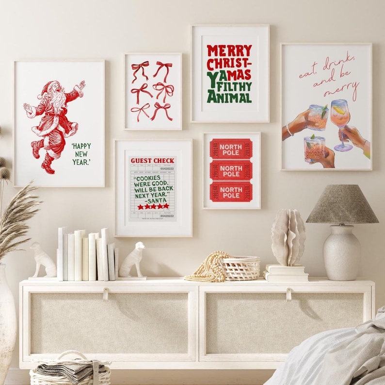 May include: A collection of six framed prints with Christmas-themed designs. The prints feature red and green text, illustrations of Santa Claus, bows, and cocktails. The prints are arranged on a white wall above a white dresser with a white lamp and a white basket.