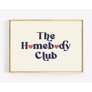 May include: A framed print with a cream background and gold frame. The text "The Homebody Club" is in navy blue with two red hearts.