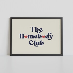 May include: A black framed print with a white background and the text "The Homebody Club" in navy blue with two red hearts.