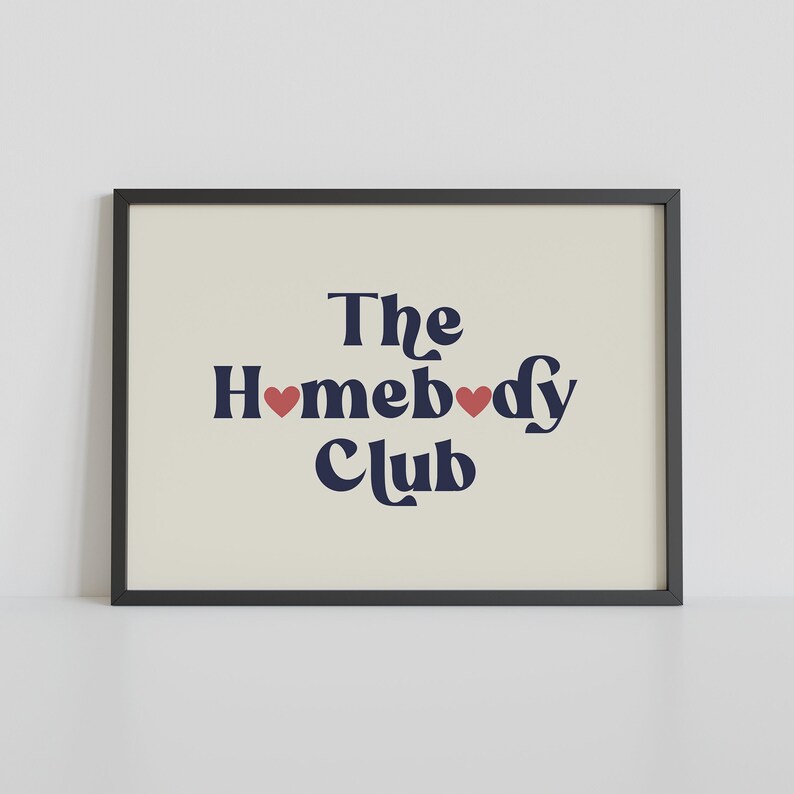 May include: A black framed print with a white background and the text "The Homebody Club" in navy blue with two red hearts.