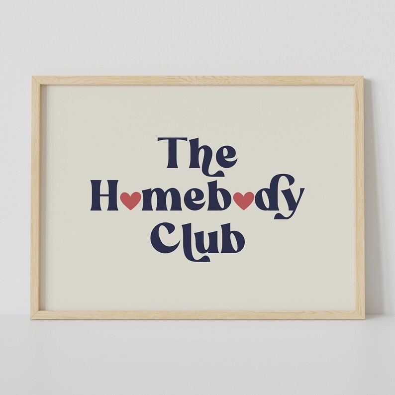 May include: A framed print with a beige background and navy blue text that reads "The Homebody Club" with two red hearts in the text.
