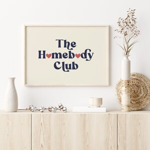 May include: A framed print with a cream background and navy blue text that reads "The Homebody Club" with two red hearts in the text. The frame is light wood and the print is sitting on a light wood shelf.