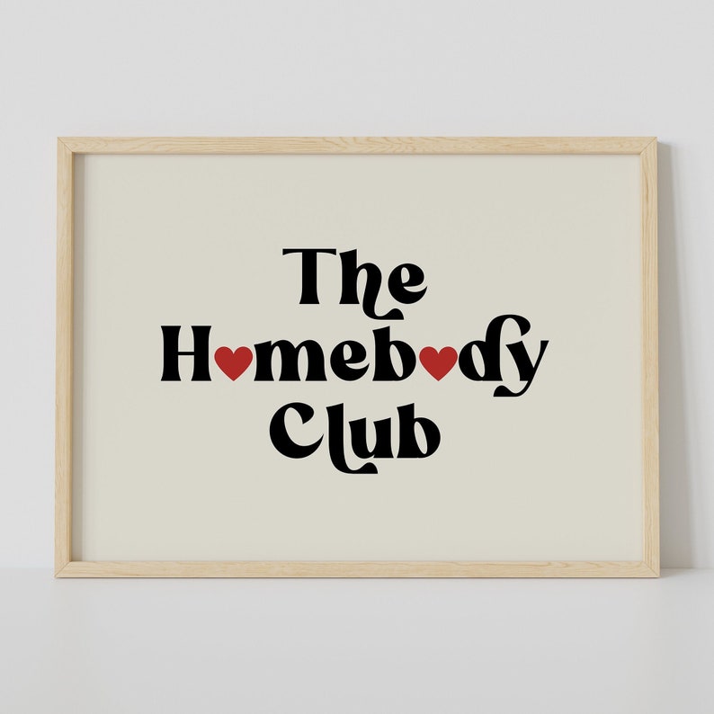 May include: A framed print with the text "The Homebody Club" in black with two red hearts. The background is a light beige.