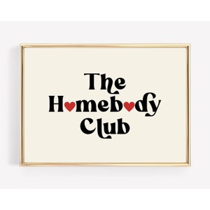 May include: A gold framed print with a white background and black text that reads "The Homebody Club" with two red hearts.