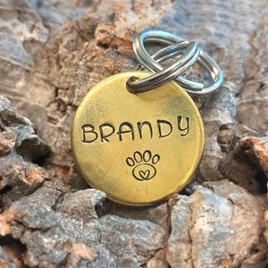 Könnte beinhalten: Ein goldfarbener Hundemarkenanhänger aus Metall mit dem eingravierten Namen "BRANDY". Der Anhänger zeigt außerdem einen Pfotenabdruck mit einem Herz darin.