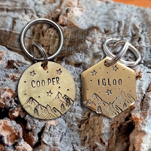 Puede incluir: Dos placas de identificación de latón con anillas plateadas. Una placa es circular y tiene grabado el nombre COOPER, mientras que la otra es hexagonal y tiene el nombre IGLOO. Ambas presentan un diseño grabado de montañas y estrellas.