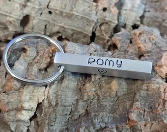 Könnte beinhalten: Ein silberfarbener Schlüsselanhänger aus Metall mit einem rechteckigen Stab, auf dessen Vorderseite der Name ROMY in schwarzen Großbuchstaben eingeprägt ist, ergänzt durch ein kleines Herzsymbol direkt unter dem Namen auf derselben Seite.