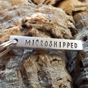 Op de afbeelding: Een zilveren sleutelhanger met een rechthoekig label waarop "MICROSHIPPED" staat.