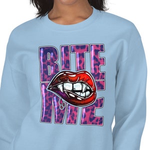 Puede incluir: Sudadera azul claro con un diseño de estampado de leopardo rosa y morado. El diseño presenta un labio rojo abierto, de tipo vampiro, con colmillos y las palabras "BITE ME" en blanco.