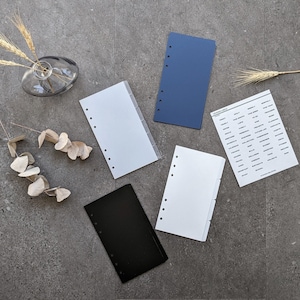 May include: Five different coloured and sized paper inserts for a personal planner. The inserts are white, black, blue, and white with black dividers. The inserts have holes punched along the left side. The inserts are on a grey surface.