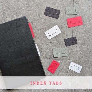 May include: A black notebook with red and white index tabs. Several rectangular index tabs in grey, red, white, and black are scattered around. The image includes the text "INDEX TABS" at the bottom.