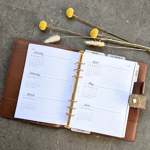 Peut inclure: Un agenda relié en cuir marron avec des anneaux dorés et des pages blanches. L'agenda comporte des onglets pour "Aujourd'hui", "Ce mois-ci" et "À faire". Les pages comprennent des calendriers mensuels de janvier à juin 2024.