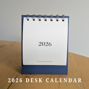May include: A navy blue desk calendar for the year 2026. The calendar has a white paper with the year printed in black, and a spiral binding. The base is a darker blue, and the text "2026 DESK CALENDAR" is at the bottom.