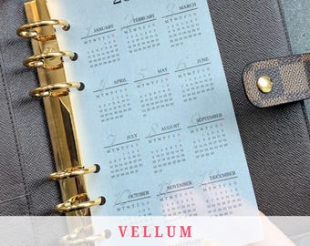 May include: A translucent vellum calendar sheet for the year 2027, featuring twelve monthly grids with black text. The sheet is held by a gold-colored six-ring metal binder mechanism inside a dark brown textured leather planner cover.