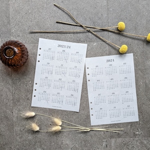 Può includere: Due pagine bianche con testo nero che mostrano un calendario per gli anni 2023-2024 e 2024. Le pagine sono su una superficie grigia con fiori secchi e un vaso marrone.