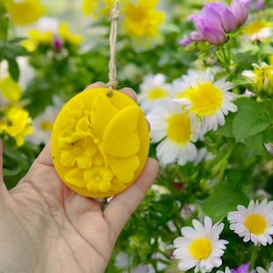 Könnte beinhalten: Ein gelber Bienenwachs-Anhänger in Form eines Schmetterlings mit Blumen. Der Anhänger hängt an einer Schnur und befindet sich vor einem Hintergrund aus weißen und gelben Blumen.