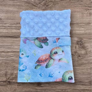 Op de afbeelding: Een babydeken met een lichtblauwe minky bovenkant en een bedrukte onderkant met cartoon zeeschildpadden, zeepaardjes en vissen in blauwe, groene en bruine tinten. De deken heeft een zachte, getextureerde uitstraling.