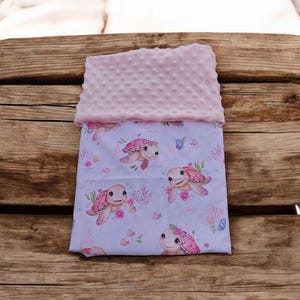 Peut inclure: Une couverture pour bébé avec un dessus rose clair texturé et un dessous imprimé. Le dessous présente un motif de tortues de mer souriantes avec des accents floraux roses, du corail et de petits poissons sur un fond bleu clair.