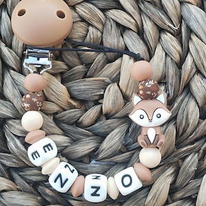 Può includere: Un mordedor per neonati in silicone marrone e bianco con un ciondolo a forma di volpe e il nome "ENZO" scritto in lettere bianche. Il mordedor è attaccato a un clip marrone e bianco.