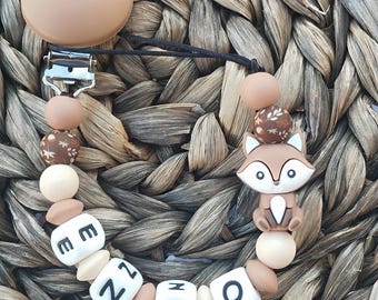 May include: A dummy clip featuring a brown silicone dummy holder, a metal clip, and a string of beads in shades of brown, beige, and white. The beads include a fox-shaped charm and white cube beads spelling the name ENZO in black letters.