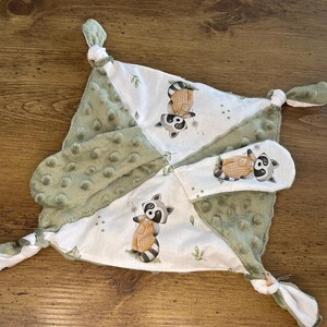 Peut inclure: Une couverture de sécurité pour bébé avec un motif blanc et vert et une impression de raton laveur. La couverture a un dos vert doux et texturé et des coins noués. Les illustrations de ratons laveurs portent des salopettes.