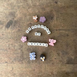 Könnte beinhalten: Eine Anordnung kleiner Dekorationsartikel auf einer Holzoberfläche. Dazu gehören ein Fuchs, eine lila Blume, eine rosa Blume, ein rosa Elefant, ein blauer Bär und ein Pinguin. Der Text "CREAZIONE DI LETIZIA" ist mit Buchstabenwürfeln geschrieben.
