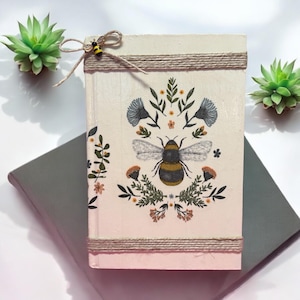 May include: A decorative journal with a bee and floral design. The cover is cream-colored with a bee illustration in the center, surrounded by leaves and flowers. The journal is tied with twine and a small bee charm. The journal is resting on top of a gray book.