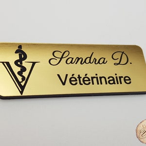 Peut inclure: Un badge rectangulaire doré avec des coins arrondis. Le badge présente un symbole de caducée noir au-dessus d'un « V » stylisé. Le nom « Sandra D. » et le mot « Vétérinaire » sont imprimés en script noir.