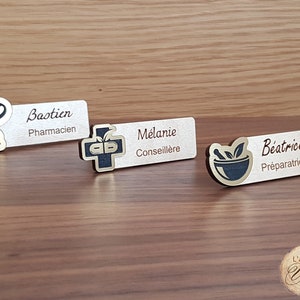 Peut inclure: Trois badges nominatifs en bois avec des détails noirs et dorés. Les badges présentent des symboles liés à la pharmacie et les noms : "Bastien, Pharmacien", "Mélanie, Conseillère", et "Béatrice, Préparatrice". Les badges sont posés sur une surface en bois.