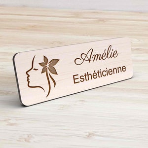 Peut inclure: Un badge en bois clair avec le nom "Amélie" et le titre "Esthéticienne" gravés en marron. Le badge présente un profil de visage de femme avec un motif floral. Le badge est rectangulaire avec des coins arrondis.