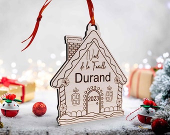Peut inclure: Ornement de Noël en bois en forme de maison en pain d'épices. L'ornement porte l'inscription gravée "Noël de la Famille Durand 2023". Il est suspendu par un ruban rouge et entouré de décorations festives.