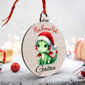 Peut inclure: Ornement de Noël en bois rond avec un dragon vert portant un bonnet de Père Noël. Le texte "Mon Premier Noel" et "Gaëtan" en rouge, avec l'année "2024" au-dessus. Un ruban rouge est attaché pour l'accrocher.