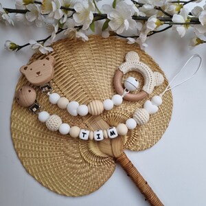 May include: A wooden baby teether with a bear head, a white crocheted ring with a bell, and white and wooden beads. The beads spell out the name "TIM".