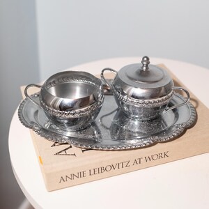 May include: A silver-colored creamer and sugar bowl set with a tray. The creamer and sugar bowl have a rope-like design around the rim. The tray has a scalloped edge and a floral pattern. The set is on top of a book that says "ANNIE LEIBOVITZ AT WORK."