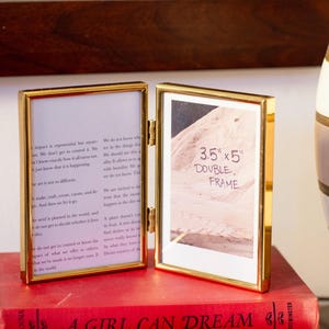 May include: A gold-colored double picture frame, open to display two images. The frame is labeled "3.5" x 5" DOUBLE FRAME". The frame is resting on a stack of red books. The frame is hinged in the middle.