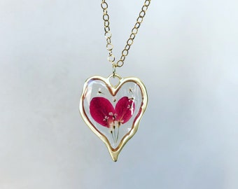 May include: A gold chain necklace with a heart-shaped pendant. The pendant is filled with clear resin and contains pressed pink flower petals.