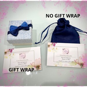 May include: Two gift wrapping options for a small item. The first option is a blue gift bag with a drawstring closure. The second option is a small white box with a blue bow. Both options have a card with the text "Ciurihandmade" and "Fiori da Indossare" in Italian and "Flowers to Wear" in English. The card also says "Gioielli in resina e fiori veri" in Italian and "Jewelry in resin and real flowers" in English. The text "NO GIFT WRAP" is written in black on a white background above the blue gift bag. The text "GIFT WRAP" is written in black on a white background below the white box.