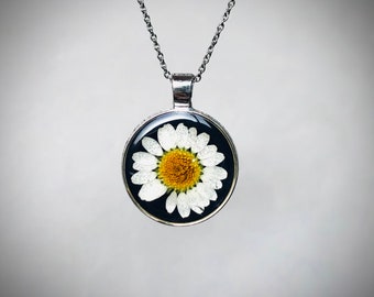 May include: A silver chain necklace with a round pendant featuring a white daisy pressed inside clear resin against a black background.
