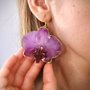 May include: Purple orchid flower earrings with gold-colored trim. The earrings are a unique floral design, showcasing the natural beauty of the orchid. The earrings are a dangle style, with a gold-colored hook.