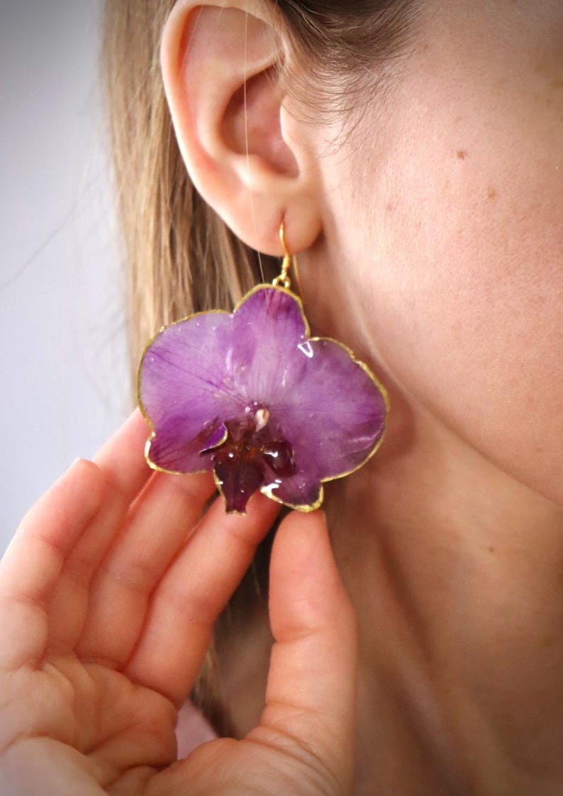 May include: Purple orchid flower earrings with gold-colored trim. The earrings are a unique floral design, showcasing the natural beauty of the orchid. The earrings are a dangle style, with a gold-colored hook.