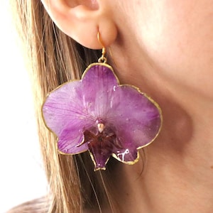 May include: A single, large, purple orchid flower earring with a gold-colored trim. The earring is a realistic representation of the flower, with detailed petals and a delicate appearance. The earring is suspended from a small gold-colored hook.