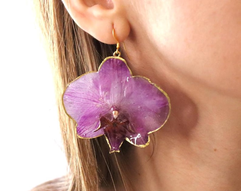 May include: A single, large, purple orchid flower earring with a gold-colored trim. The earring is a realistic representation of the flower, with detailed petals and a delicate appearance. The earring is suspended from a small gold-colored hook.