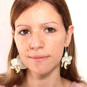 May include: A woman with long brown hair wearing white flower earrings. The earrings are made of white flowers with gold accents.